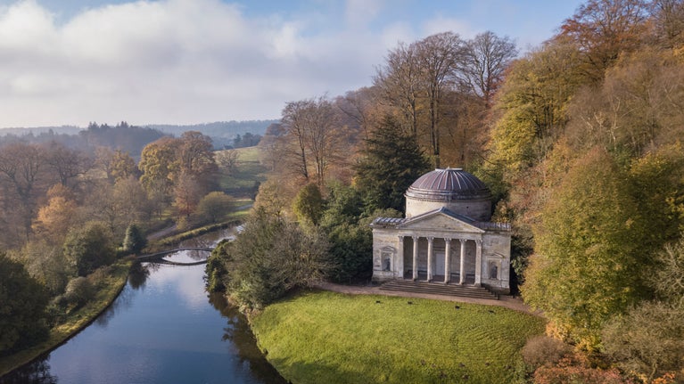View of the Pantheon and lake in October at Stourhead, Wiltshire with autumnal trees in the foreground and a blue sky with clouds